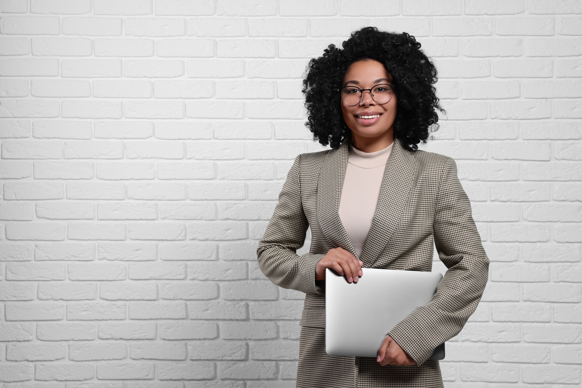 Young businesswoman holding laptop near white brick wall. Space for text